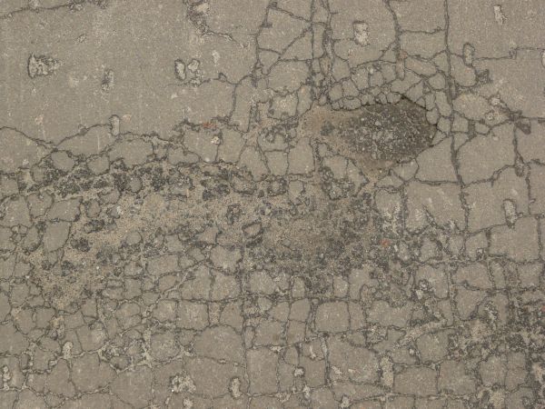 Grey asphalt texture covered with thick cracks and with several small broken holes full of dirt and rocks.